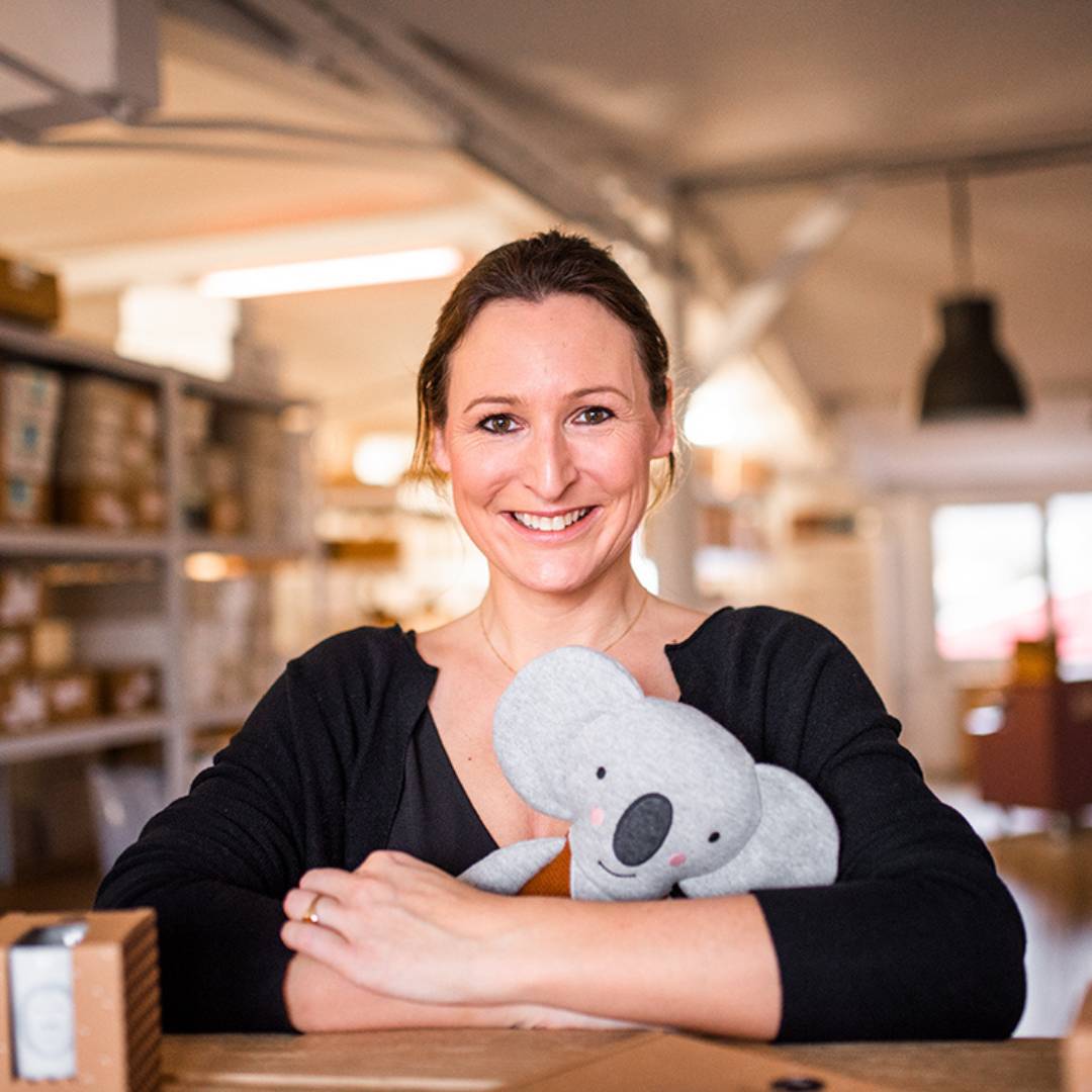 Geraldine, die Gründerin von ava&yves hält sitzend hinter einem Tisch ein Kuscheltier in der Hand und lächelt in die Kamera